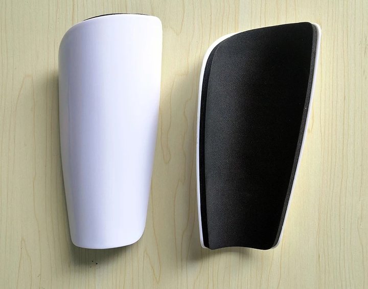 Plain white full-size shin guard, front face and foam-lined back, on a wooden surface