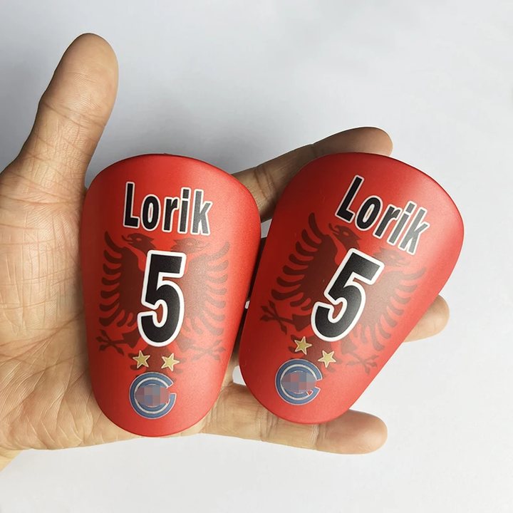 Pair of red mini shin guards printed 'Lorik 5' held in a hand for scale